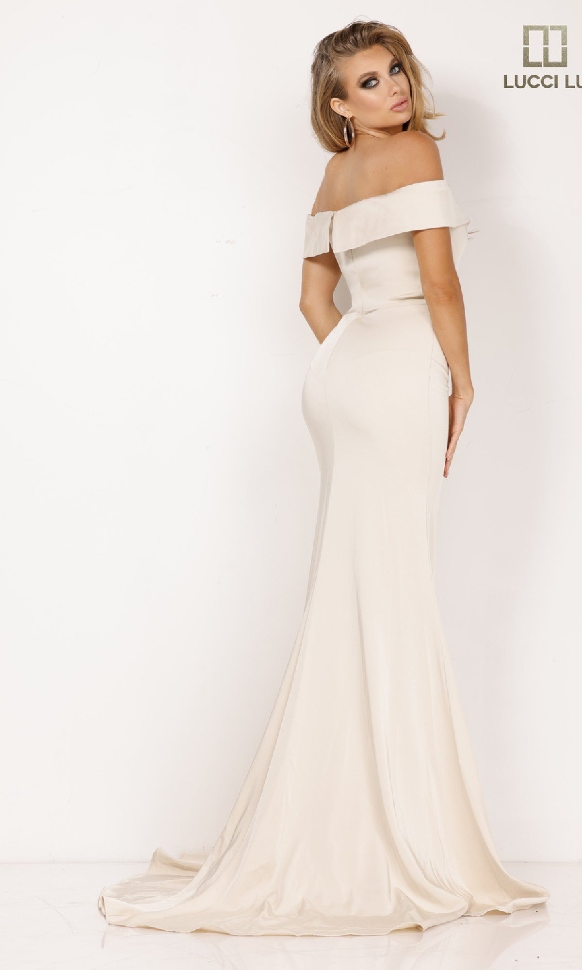 A woman poses gracefully against a plain white background in the Lucci Lu 8262 Off-the-Shoulder Crepe Prom Dress, a floor-length fitted cream gown with a flared train—an elegant choice for formal occasions.